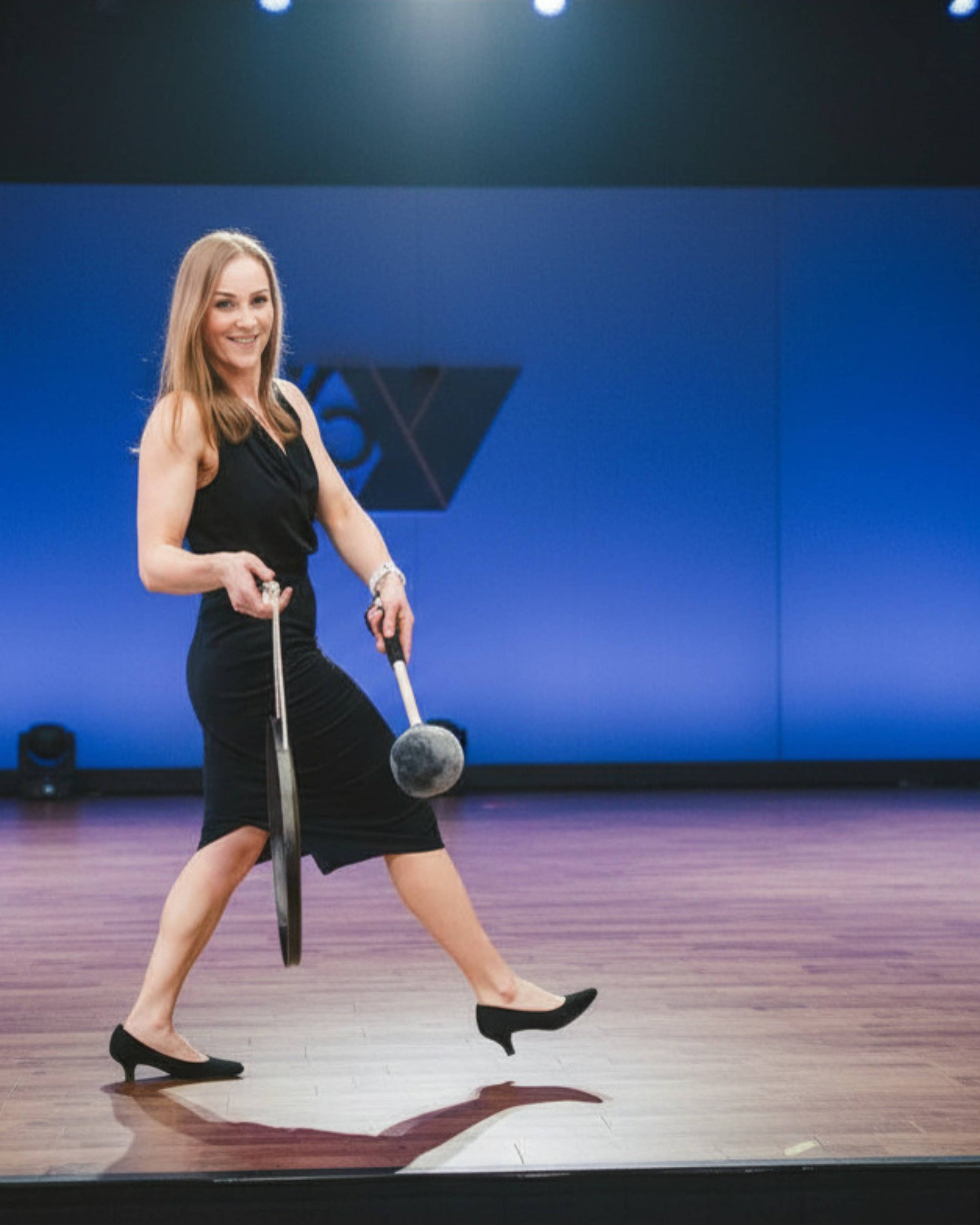 Liz Adriano walking onstage to speak to a corporate convention audience in a black dress holding a gong on a stage with a blue event background.