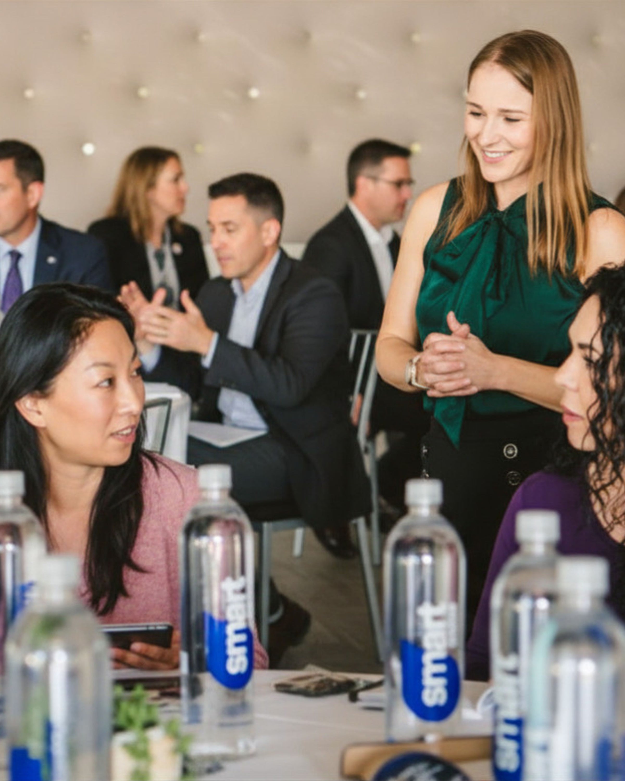 Liz Adriano facilitating and engaging with attendees at a corporate event picturing also groups of people at a conference table with water bottles in front.
