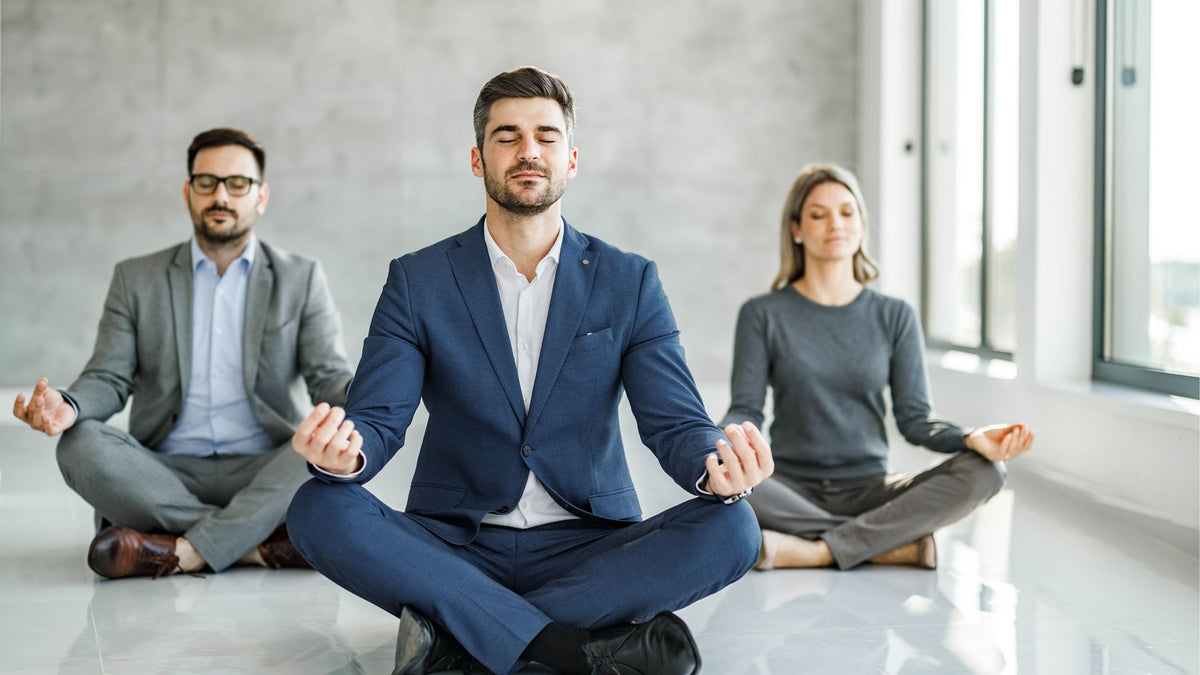 Three executives in business attire sitting in a meditation position on a light-colored conference room floor during an executive workshop.