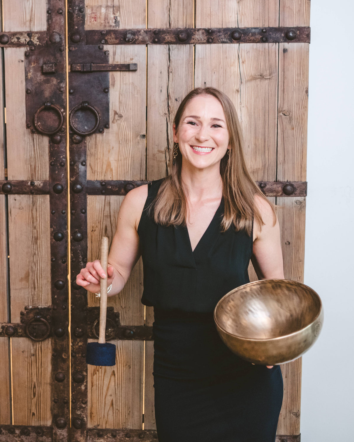 Ascendify Wellness Founder and Proprietary Event Designer, Liz Adriano, holding a bronze singing bowl and mallet in front of a wooden door.