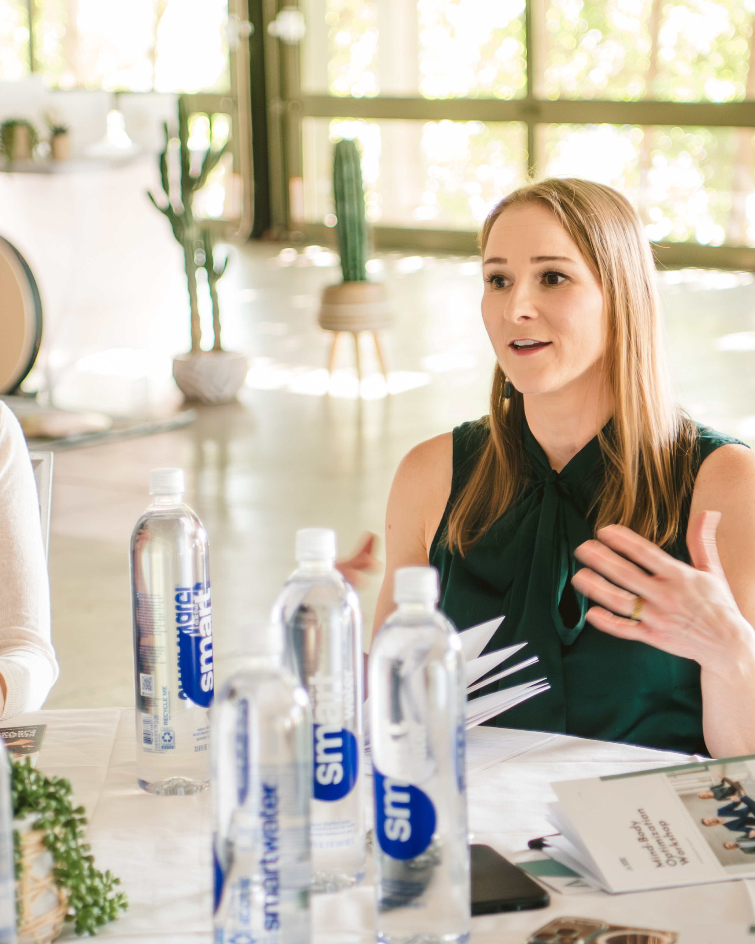 Liz Adriano, interactive keynote speaker, speaking at a table at a corporate event with water bottles and custom workbooks in a bright event venue.