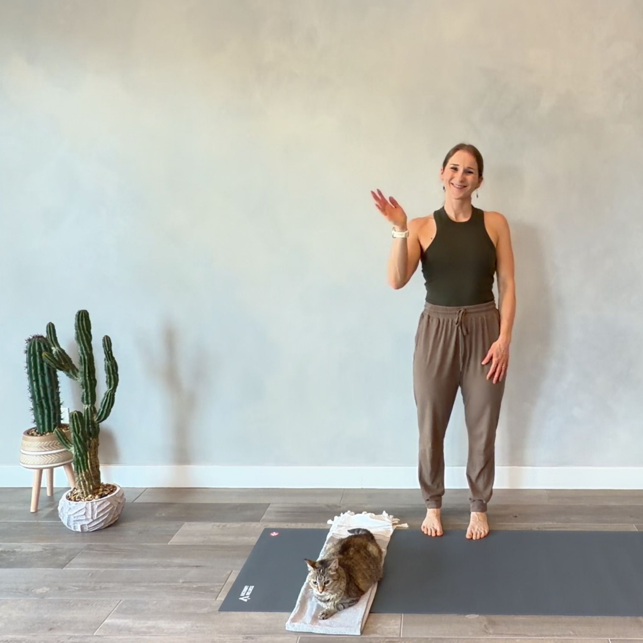 Woman in a yoga studio waving with a cat on a mat with a cactus plant in the background.